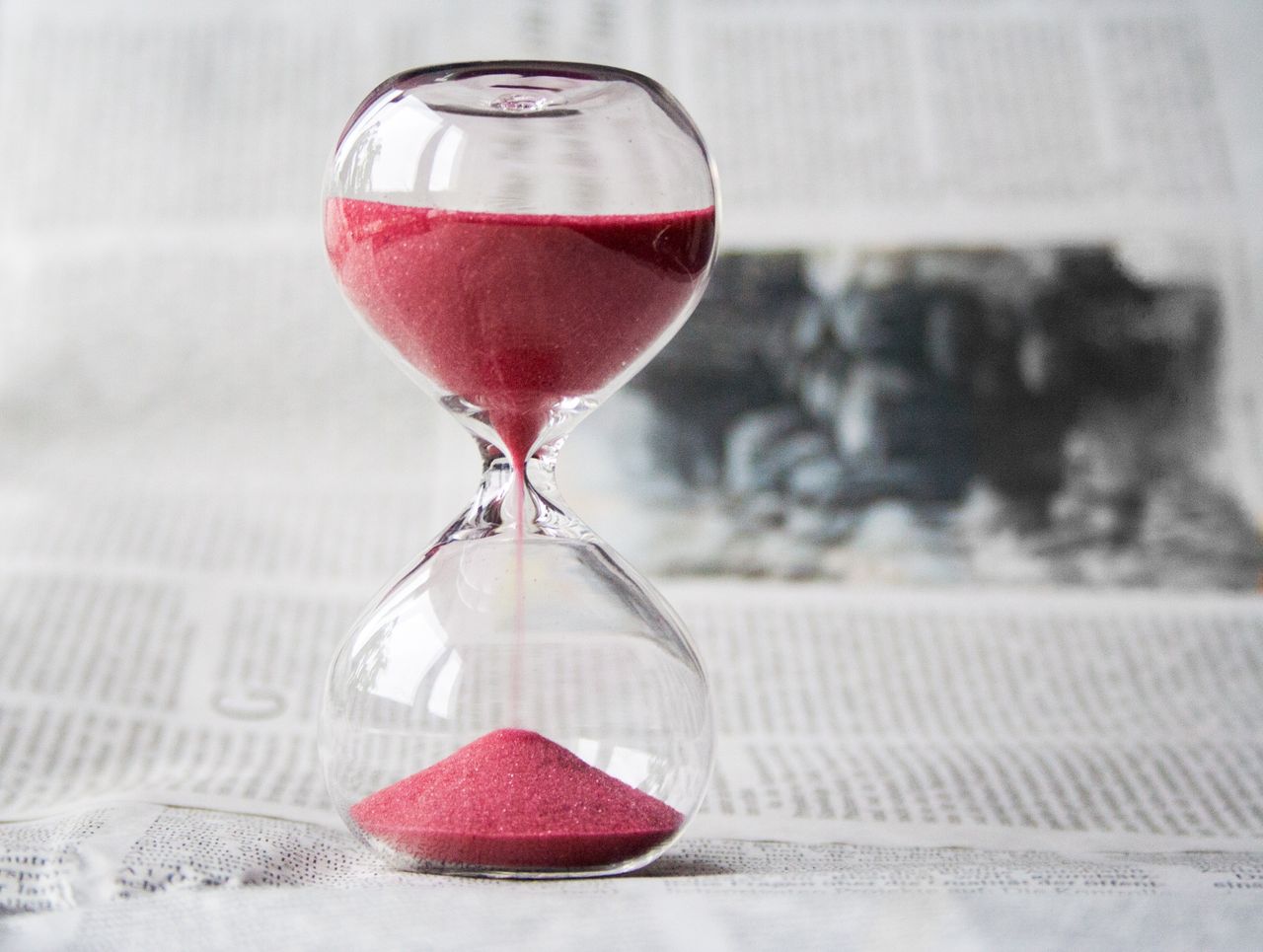 about-02 Close-up of a transparent hourglass with pink sand flowing, placed on a newspaper background.